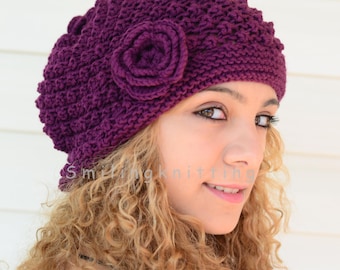 May include: A purple knitted beanie with a large flower detail. The beanie is worn by a person with curly blonde hair.