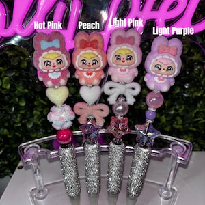 May include: Four decorative pens in pink, peach, light pink, and light purple. Each pen has a rhinestone-covered barrel and a unique topper with a cartoon character in a bunny costume. The pens are displayed in a clear plastic holder.