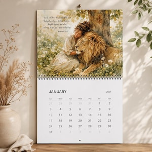 May include: A 2027 wall calendar featuring a January page with a printed illustration of a person in a light-coloured dress embracing a large lion in a field of white flowers. The text Jeremiah 29:11 is visible in the upper left corner of the image.