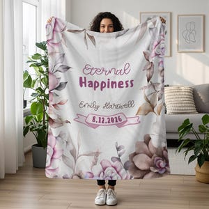 May include: A white blanket with a floral design in shades of pink, purple, and brown. The blanket features the text "Eternal Happiness," a name, and a date. The design includes watercolor-style flowers and leaves.