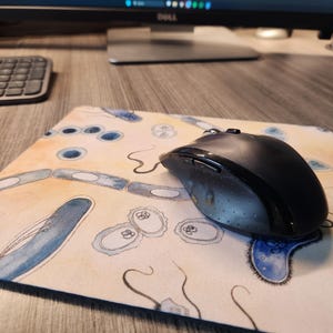 Pode incluir: Um rato de computador sem fios preto repousa sobre um tapete de rato bege. O tapete do rato apresenta um desenho em aguarela de formas biológicas azuis e bege. Um monitor de computador e um teclado são visíveis no fundo.