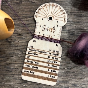 May include: A wooden yarn gauge tool with the text "Smash Knits" and measurements in inches and centimeters. The gauge tool is used to measure yarn weight. The image also shows yarn and fiber.