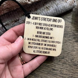 May include: A wooden tag with the text "JENNY'S STRETCHY BIND OFF" and knitting instructions. The tag is attached to a knitted item with a black cord. The background is a wooden surface.