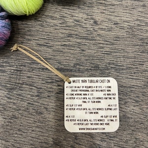 May include: A close-up of a wooden tag with instructions for a "Waste Yarn Tubular Cast On" knitting technique. The tag is attached to a length of twine and is sitting on a wooden surface.