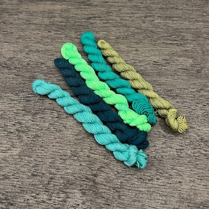 May include: Six skeins of embroidery floss in shades of green, blue, and yellow. The floss is arranged in a fan shape.