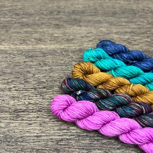 May include: Five skeins of yarn in different colors, including pink, teal, gold, green and blue. The yarn is arranged in a fan shape on a wooden surface.