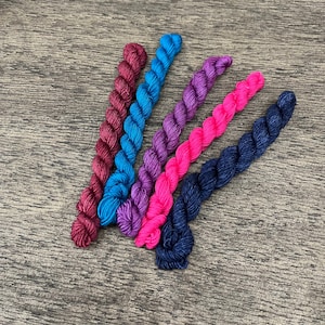 May include: Five skeins of yarn in different colors: burgundy, teal, purple, hot pink, and navy blue. The yarn is arranged in a fan shape on a wooden surface.