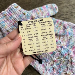 May include: Hand-knitted mittens in pastel shades, held with a mitten size chart tag. The tag provides measurements in inches and cm for various sizes, including child, adult, and XL. The website address www.smashknits.com is visible.