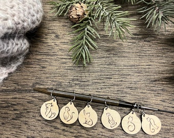May include: Set of six wooden cat stitch markers with black metal rings. The markers are round and feature a simple line drawing of a cat on each one. The markers are arranged in a row on a wooden surface.