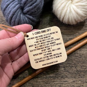 May include: A wooden tag with instructions for an I-cord bind off, held near two balls of yarn and knitting needles. The tag has a leather loop and text that reads "I-CORD BIND OFF" and instructions. The yarn is gray and cream colored.