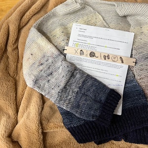 May include: A gray and blue knitted sweater with a white and gray ombre effect. The sweater is lying on a brown fuzzy blanket. A printed sheet of paper with the title "Knit Card" is partially visible under the sweater. The paper has a list of instructions and a wooden ruler with knitting symbols.