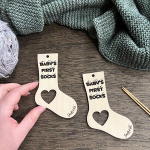 Könnte beinhalten: Zwei hölzerne Babysocken-Ornamente mit Herz-Ausschnitten. Der Text "BABY'S FIRST SOCKS" ist auf jedem Ornament aufgedruckt. Eine Wollkugel, Stricknadeln und eine Strickdecke befinden sich im Hintergrund.