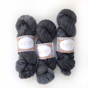 Peut inclure: Trois écheveaux de fil gris foncé avec un dégradé de gris plus clair. Chaque écheveau porte une étiquette indiquant "Smush Knits Fibre Goods".