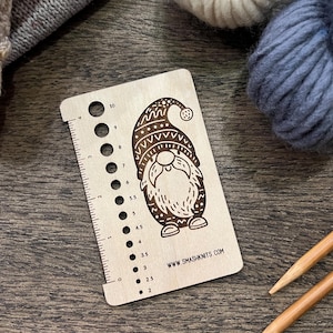May include: A wooden knitting gauge featuring a gnome illustration. The gauge has numbered holes for needle sizes and a ruler. The gnome wears a patterned hat. The website address www.smashknits.com is printed on the gauge. Knitting needles and yarn are in the background.