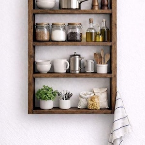 May include: A four-tier dark wood wall shelf holding various kitchen items including ceramic bowls, a stainless steel saucepan, wooden pepper mills, glass storage jars with lids, oil bottles, a metal coffee press, mugs, a plant, and utensils.