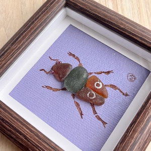Puede incluir: Obra de arte enmarcada que presenta un escarabajo de vidrio marino sobre un fondo lavanda. El escarabajo está compuesto por piezas de vidrio marino marrón, verde y naranja. El marco es de madera marrón oscuro con un paspartú blanco. Una pieza única, hecha a mano.