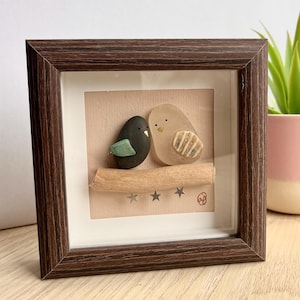 Puede incluir: Una obra de arte enmarcada con dos pájaros de guijarros posados en una ramita. Uno es negro con un ala verde, el otro es beige con detalles a rayas. El fondo es marrón claro con estrellas.