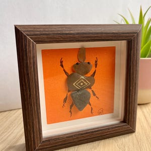 Könnte beinhalten: Gerahmtes Kunstwerk mit einem Stein-Käfer auf orangefarbenem Hintergrund. Der Käfer besteht aus verschiedenen Steinen, mit einem goldenen Diamantmuster auf seinem Körper. Der Rahmen ist dunkelbraun und das Kunstwerk wird auf einer Holzoberfläche präsentiert.