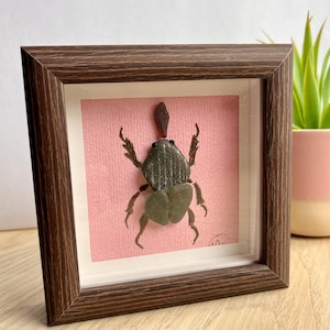 Pode incluir: Uma obra de arte emoldurada de insetos. A obra apresenta um besouro verde escuro detalhado feito de materiais naturais, contra um fundo rosa. A moldura é de madeira marrom escura e a peça é exibida em uma caixa de sombra.