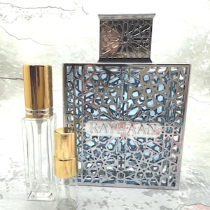 May include: A perfume bottle set featuring a large, silver-colored bottle with a geometric design and the word "RAYHAAN". Two smaller, clear glass bottles with gold-colored caps are in front of the larger bottle. The set is on a light-colored surface.