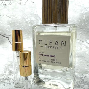 May include: A clear glass perfume bottle with a wooden cap and the text "CLEAN RESERVE". Two smaller glass spray bottles with gold caps are in front of the larger bottle. The label reads "skin [reserve blend] eau de parfum floral musk".