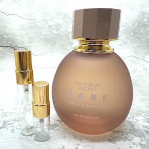 May include: A perfume bottle and two small spray bottles. The large bottle is round with a brown cap and gold accents. The text "Victoria's Secret BARE Sueded Vanilla Eau de Parfum" is printed on the bottle. The small bottles have gold caps.
