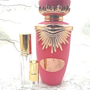 May include: A large pink perfume bottle with a textured matte finish, featuring a gold-toned metallic emblem shaped like an eye with a small red stone, paired with two smaller clear glass spray vials with gold caps standing in the foreground.