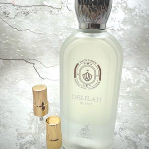 May include: A perfume bottle with a silver cap and the words "DELILAH BLANC" printed on the front. Two small, empty spray bottles with gold caps are in front of the perfume bottle. The perfume bottle has a frosted appearance.