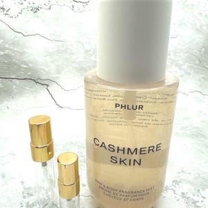 May include: A clear, cylindrical bottle of PHLUR Cashmere Skin fragrance mist with a white cap. Two small, empty spray bottles with gold caps are next to it. The text "Cashmere Skin" is visible on the bottle.