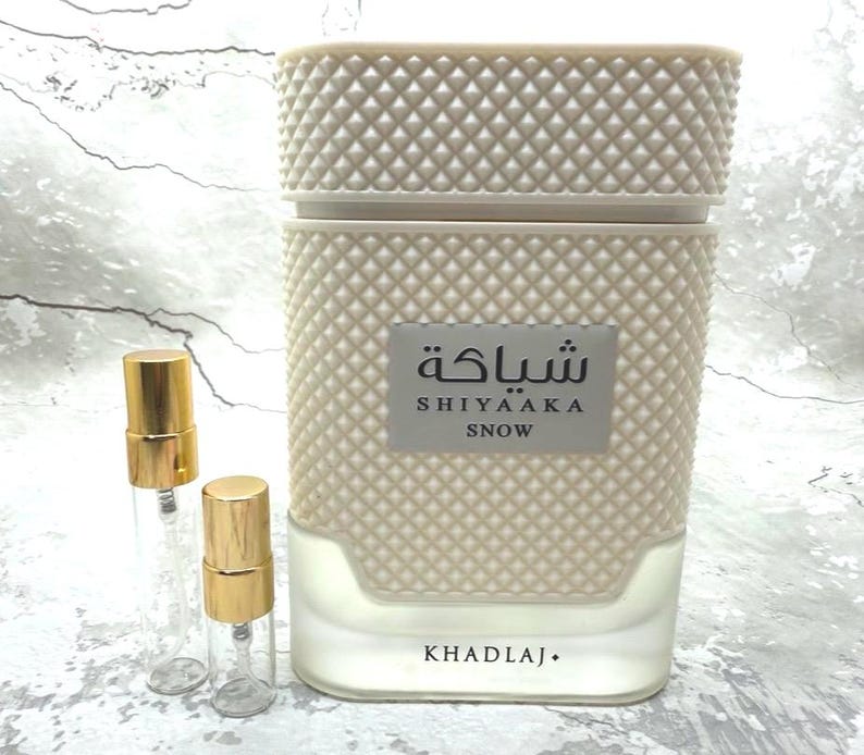 Puede incluir: Un frasco de perfume beige con un patr&oacute;n de diamantes texturizado, etiquetado como "SHIYAAKA SNOW" en ingl&eacute;s y &aacute;rabe. Dos peque&ntilde;os viales de vidrio transparente con tapas doradas est&aacute;n junto al frasco. La base del frasco dice "KHADLAJ."