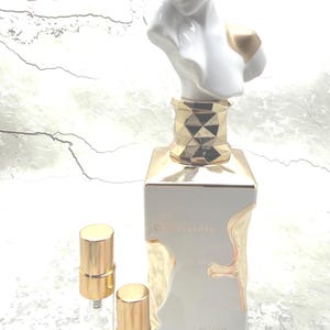 May include: A white and gold perfume bottle featuring a sculpted bust topper with gold accents on the eyes and shoulder. The square base has a gold geometric neck and gold drip-like detailing. Two small clear glass vials with gold caps sit nearby.