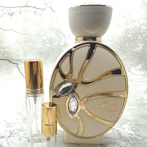 May include: A perfume bottle with a cream-colored base and gold accents. The bottle has a large, round design with a gold pattern and two clear, gem-like embellishments. The text "Khadlaj" is visible on the top. Two smaller, clear bottles with gold caps are in front.