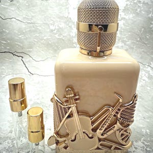 May include: A beige perfume bottle with a gold microphone-shaped cap and musical instrument embellishments. Two small, empty spray bottles with gold caps are next to the perfume bottle. The text "FRAGRANCE WORLD" is at the bottom.