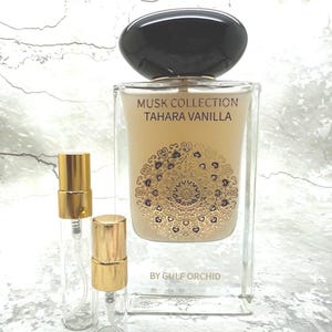 May include: A rectangular perfume bottle with a black cap and gold accents. The bottle is labeled "Musk Collection Tahara Vanilla" with a decorative gold design. Two small, clear spray bottles with gold caps are in front of the perfume bottle.