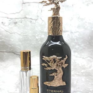 May include: A black perfume bottle with a gold tree design and a gold tree-shaped cap. The bottle is labeled "Eternal Vanille" by Lattafa Pride. Two smaller, clear bottles with gold caps are in front.