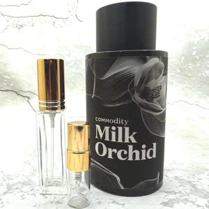 May include: A black cylindrical bottle with the text "commodity Milk Orchid" and a white orchid design, accompanied by two clear glass perfume bottles with gold caps. The bottles are of different sizes.