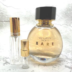 May include: A perfume bottle with a black cap and gold accents, labeled "Victoria's Secret BARE." Two smaller, clear glass spray bottles with gold caps are also present. The perfume bottle has a rounded shape.