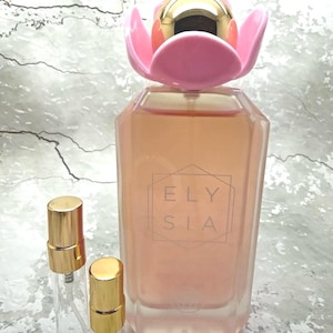 May include: A perfume bottle with a pink flower-shaped cap and a gold top. The bottle is filled with a light peach-colored liquid and has the word "ELYSIA" printed on the front. Two small glass spray bottles with gold caps are next to the perfume bottle.