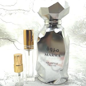 May include: A silver perfume bottle with a geometric design, accompanied by two smaller clear glass spray bottles with gold-colored caps. The large bottle has the word "MARWA" and "ARABIYAT PRESTIGE" printed on it.