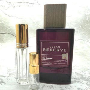 May include: A collection of perfume bottles. The largest bottle is a deep burgundy with a wooden cap and a label that reads "CLEAN RESERVE". Two smaller, clear glass bottles with gold-colored caps are in front.