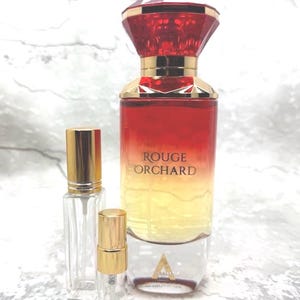 May include: A perfume bottle with a red and yellow gradient, gold accents, and a faceted red cap. The bottle is labeled "ROUGE ORCHARD." Two smaller, clear glass bottles with gold caps are also present.
