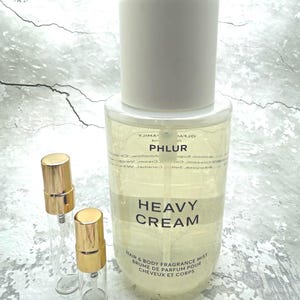 May include: A clear glass bottle of "HEAVY CREAM" hair and body fragrance mist with a white cap. Two small glass vials with gold-colored caps are next to the bottle. The text "PHLUR" is visible on the bottle.