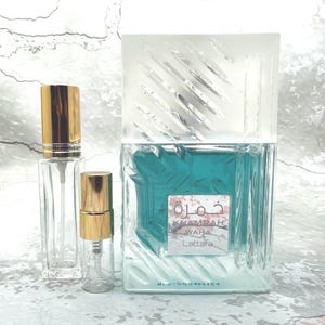 May include: A clear glass perfume bottle with a gold cap stands next to two smaller clear glass spray bottles with gold caps. The largest bottle has a square shape with a turquoise liquid inside and a frosted glass top. The label reads "Khamrah Waha Lattafa."