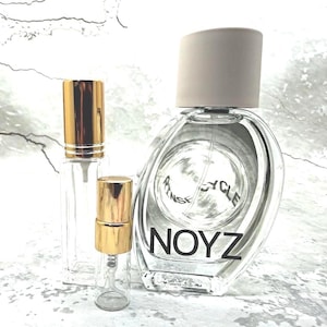 May include: A large oval glass perfume bottle with a beige cap and the text NOYZ printed in black, accompanied by two smaller clear glass spray vials featuring shiny gold caps and clear plastic spray mechanisms for fragrance storage.