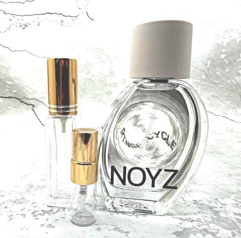 May include: A large oval glass perfume bottle with a beige cap and the text NOYZ printed in black, accompanied by two smaller clear glass spray vials featuring shiny gold caps and clear plastic spray mechanisms for fragrance storage.