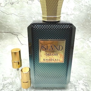May include: A perfume bottle with a gold cap and a textured, dark teal body. The bottle has a gold label that reads "ISLAND DREAMS KHADLAJ." Two small, clear spray bottles with gold caps are in front of the perfume bottle.