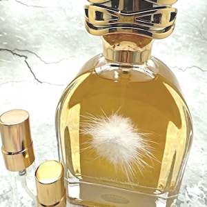 May include: A gold-coloured perfume bottle with a decorative gold cap and a fluffy white design on the front. Two small, clear spray bottles with gold caps are next to the perfume bottle. The perfume bottle is filled with a golden liquid.