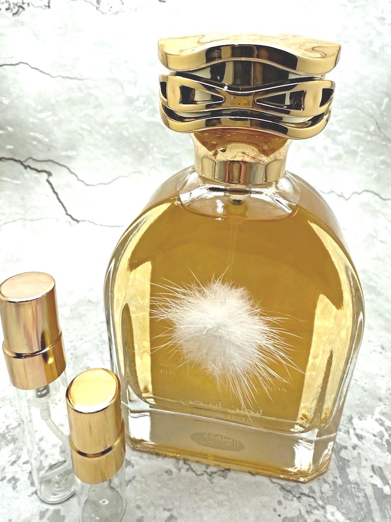 May include: A gold-coloured perfume bottle with a decorative gold cap and a fluffy white design on the front. Two small, clear spray bottles with gold caps are next to the perfume bottle. The perfume bottle is filled with a golden liquid.