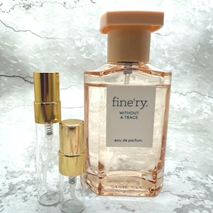 May include: A perfume bottle and two travel-size spray bottles. The perfume bottle is rectangular with a peach-colored cap and contains a light pink liquid. The label reads "fine'ry. WITHOUT A TRACE eau de parfum". The travel bottles have gold-colored caps.