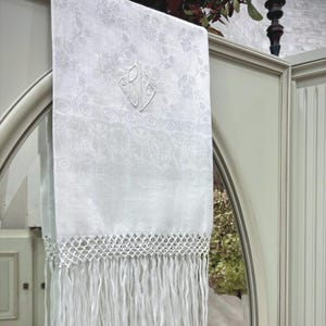 Puede incluir: Toalla de lino blanco con estampado floral y flecos. La toalla presenta un monograma bordado en forma de diamante. El borde inferior tiene un diseño calado con flecos largos.